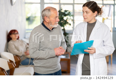 Young nurse or doctor talking with elderly man in reception area Young nurse or doctor talking with elderly man in reception area 142494129