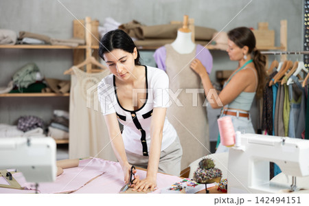 Young seamstress cutting fabric in sewing workshop with colleague Young seamstress cutting fabric in sewing workshop with colleague 142494151
