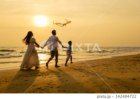 Happy family enjoying holiday vacation by flying kite together on sandy beach near ocean water during warm sunset light, creating joyful moments outdoors 142494722