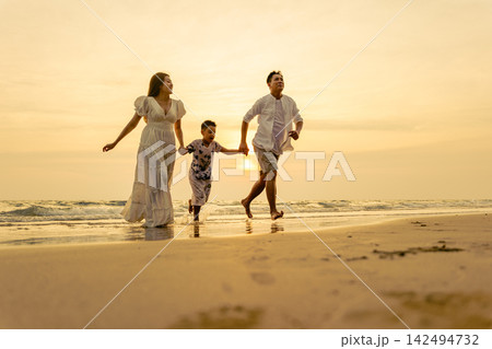 Family enjoying holiday vacation on beach at sunset, running together near ocean water while having fun during joyful seasonal trip outdoors with bright sunlight lighting landscape 142494732