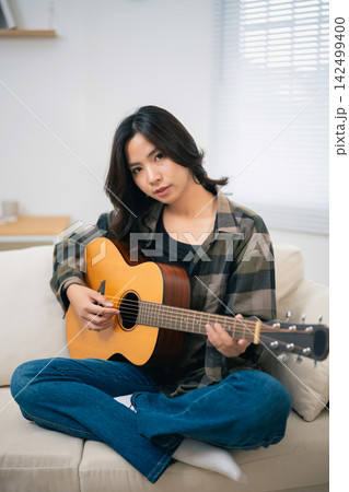 Young woman sitting cross-legged on sofa playing acoustic guitar with focused expression at home in casual outfit Young woman sitting cross-legged on sofa playing acoustic guitar with focused expression at home in casual outfit 142499400