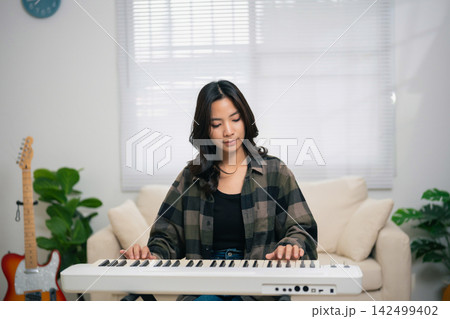 Young woman playing electronic keyboard indoors with focused expression sitting in cozy room with plants and guitar in background Young woman playing electronic keyboard indoors with focused expression sitting in cozy room with plants and guitar in background 142499402