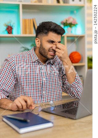 Indian man on home table rubbing tired eyes from laptop work and feeling fatigue during remote job Indian man on home table rubbing tired eyes from laptop work and feeling fatigue during remote job 142502224