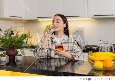 Young woman drinks water and checks news online on orange smartphone, hydration and wellness advice 142502231