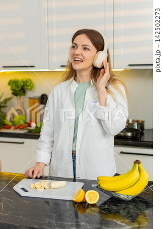 Middle-aged woman listening to music while slicing banana for dietary smoothie snack in home kitchen Middle-aged woman listening to music while slicing banana for dietary smoothie snack in home kitchen 142502273