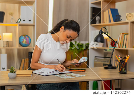 African American young woman on home table writing down expenses and counting cash for budget plan 142502274