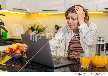 Young woman feeling stressed by laptop in home kitchen during diet break after upsetting online news Young woman feeling stressed by laptop in home kitchen during diet break after upsetting online news 142502633