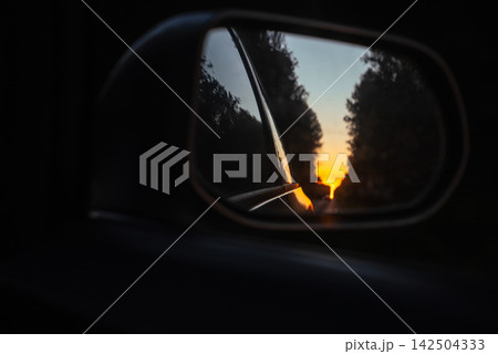 Closeup Of Sunset Reflected In Car Side Mirror On Tree Lined Road. Travel, Driving, Transportation And Road Trip Concept. Closeup Of Sunset Reflected In Car Side Mirror On Tree Lined Road. Travel, Driving, Transportation And Road Trip Concept. 142504333