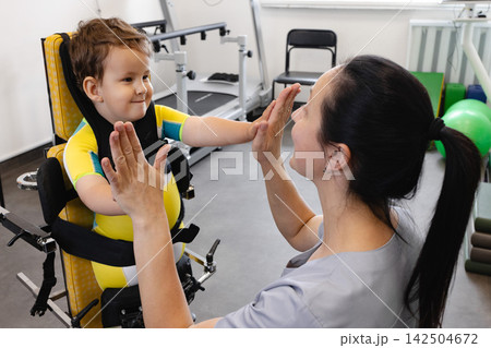 Happy boy during physical rehabilitation standing in a standing frame with his physical therapist, working on developing standing skills Happy boy during physical rehabilitation standing in a standing frame with his physical therapist, working on developing standing skills 142504672