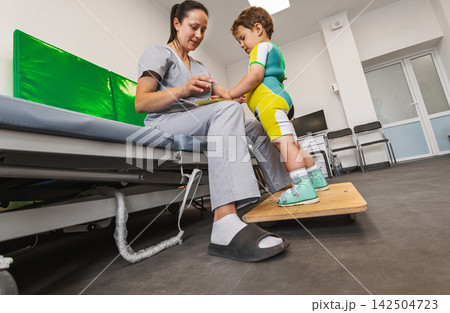 Child patient stands on balance platform while a therapist assists with rehabilitation in a clinic. Balance disorder, ataxia Child patient stands on balance platform while a therapist assists with rehabilitation in a clinic. Balance disorder, ataxia 142504723