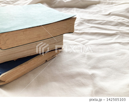 ベッドルームでの休日読書やリラックスタイム・白いシーツの上に重ねられた古本・読書習慣・ライフスタイル ベッドルームでの休日読書やリラックスタイム・白いシーツの上に重ねられた古本・読書習慣・ライフスタイル 142505103