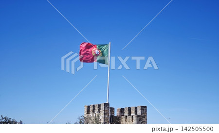 Flag of Portugal waving atop a stone castle tower against a clear blue sky, showcasing historical architecture and national pride in a scenic outdoor setting Flag of Portugal waving atop a stone castle tower against a clear blue sky, showcasing historical architecture and national pride in a scenic outdoor setting 142505500