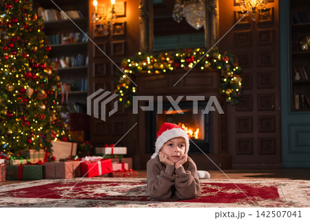 Merry Christmas. Little boy smiling near Christmas tree in classical dark interior. Young happy kid wearing Santa Hat in living room with fireplace Christmas tree gift boxes. Christmas eve at home 142507041