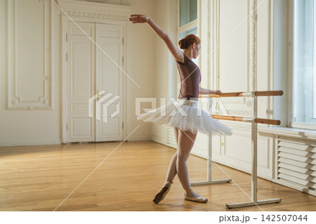 Young ballerina in tutu skirt stretching practicing ballet positions dancing near ballet barre in dance class. Young classical dancer woman in dance studio. Ballet class training Ballet school concept Young ballerina in tutu skirt stretching practicing ballet positions dancing near ballet barre in dance class. Young classical dancer woman in dance studio. Ballet class training Ballet school concept 142507044