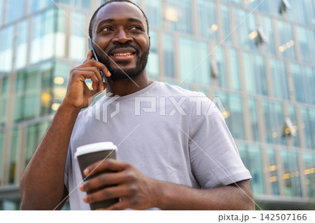 Good morning. African man drinking coffee to go talking on smartphone outdoor. Man with cell phone paper cup walking on street. Man making answering call by cellphone having conversation by mobile 142507166
