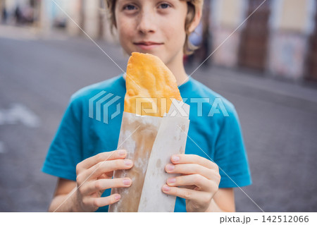 Boy holding a golden molote poblano traditional fried Mexican empanada outdoors on a street in Puebla Mexico with colonial city background paper wrapper local street food authentic travel snack. Boy holding a golden molote poblano traditional fried Mexican empanada outdoors on a street in Puebla Mexico with colonial city background paper wrapper local street food authentic travel snack. 142512066