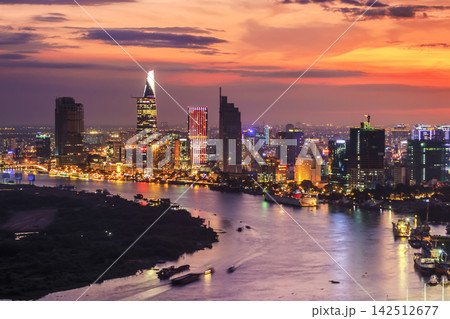 A dense modern city skyline glows beside a wide river beneath a vivid orange and purple sunset sky. A dense modern city skyline glows beside a wide river beneath a vivid orange and purple sunset sky. 142512677