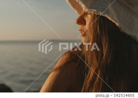 Woman in a wide brim sun hat gazes toward the calm sea at golden hour, with warm light illuminating her hair and a tranquil coastal setting creating an atmospheric summer lifestyle portrait for travel 142514605
