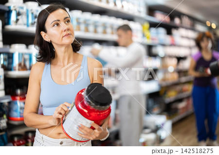 Focused woman choosing protein powder jar in sports nutrition store 142518275