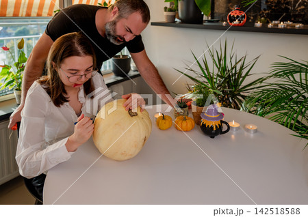 Her father leans in to help a daughter carve a pumpkin at a table decorated with mini pumpkins, candles and a bat ornament on the wall. A warm autumn scene of a father and daughter celebrating 142518588