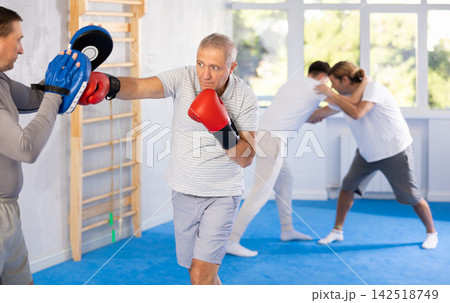 Coach and elderly man pupil are engaged during training, boxing classes. Male trainer holds makiwara and helps student to work out force of blow, to box quickly and effectively Coach and elderly man pupil are engaged during training, boxing classes. Male trainer holds makiwara and helps student to work out force of blow, to box quickly and effectively 142518749