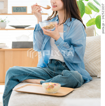 自宅のソファでシリアルを食べる女性。くつろぎながら楽しむ朝食時間 自宅のソファでシリアルを食べる女性。くつろぎながら楽しむ朝食時間 142521837