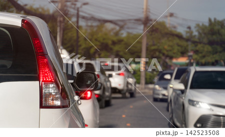 Cars wait in a traffic queue on an urban street of asphalt road. During a busy daytime commute under bright light. Blurred of trees forest side of road. 142523508