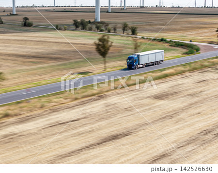 Blue semi truck carries cargo on rural road through harvested farmland, seen from drone with motion blur. Wind turbines across fields connect logistics, agriculture, transport and renewable energy Blue semi truck carries cargo on rural road through harvested farmland, seen from drone with motion blur. Wind turbines across fields connect logistics, agriculture, transport and renewable energy 142526630