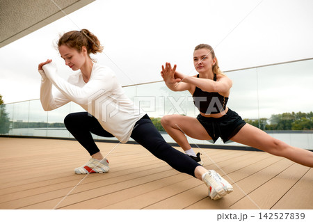 Two women doing side lunge stretch exercise on outdoor terrace Two women doing side lunge stretch exercise on outdoor terrace 142527839