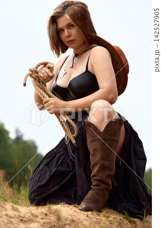 Confident woman in cowgirl attire kneeling with rope outdoors Confident woman in cowgirl attire kneeling with rope outdoors 142527905