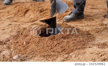 Close view shows metal auger boring into loose sandy soil while worker boots stand nearby, with auger, drilling, excavation, groundwork detail, showing construction, soil, sand, earth context 142529961