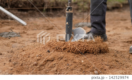 Close view of manual digging tool cutting into dry sandy ground while worker boots stand nearby, with digging, soil, sandy, ground detail, showing construction, excavation, drilling, auger context Close view of manual digging tool cutting into dry sandy ground while worker boots stand nearby, with digging, soil, sandy, ground detail, showing construction, excavation, drilling, auger context 142529974