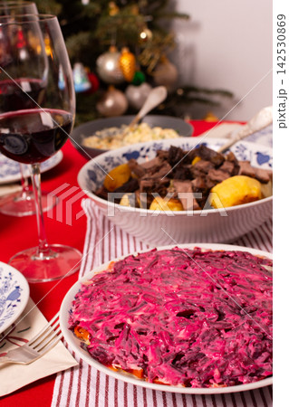 A table set for a Christmas and New Year celebration displays various dishes like salad, meat, and grains. A red candle, wine glass, and colorful decorations add to the festive atmosphere 142530869
