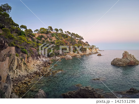 Twilight falls over the rugged rocky coastline of Lloret de Mar, Spain. Calm turquoise Mediterranean waters lap against stone outcrops below steep cliffs covered in pine trees Twilight falls over the rugged rocky coastline of Lloret de Mar, Spain. Calm turquoise Mediterranean waters lap against stone outcrops below steep cliffs covered in pine trees 142531296