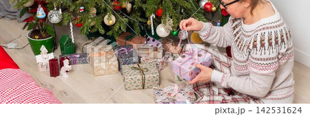 A woman aged 52 sits on the floor next to a decorated Christmas tree. She opens gifts on Christmas Day while enjoying the holiday with family members in the background A woman aged 52 sits on the floor next to a decorated Christmas tree. She opens gifts on Christmas Day while enjoying the holiday with family members in the background 142531624