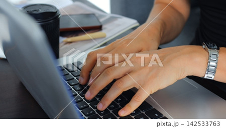 Hands typing on laptop keyboard beside coffee cup notebook and pen on desk in bright office workspace, close view of focused person working on computer for business, communication and remote 142533763