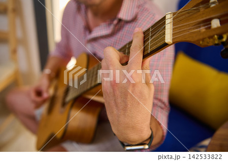 Man playing acoustic guitar at home with close-up of hand on fretboard Man playing acoustic guitar at home with close-up of hand on fretboard 142533822