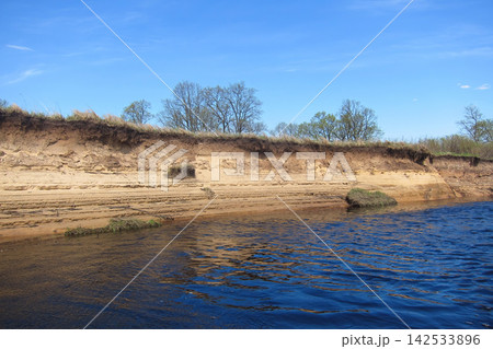 Eroded sandy riverbank with layers of soil and grass against a blue sky. Eroded sandy riverbank with layers of soil and grass against a blue sky. 142533896