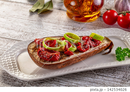 Grilled eggplant half topped with sun dried tomatoes and leek slices on white plate 142534026