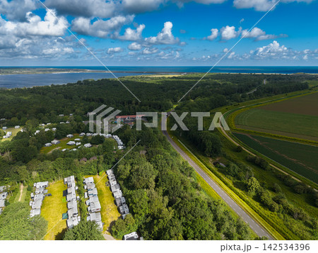 High angle wide horizontal aerial drone photograph of campsite, woodland, coastal road, grass dike, and North Sea horizon near Kamperland, Zeeland, Netherlands on sunny summer day 142534396