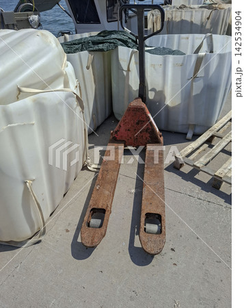 Weathered manual pallet jack parked on concrete near bulk containers on a dock 142534904