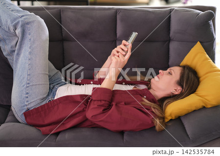 Relaxed woman uses smartphone while lying on office sofa Relaxed woman uses smartphone while lying on office sofa 142535784