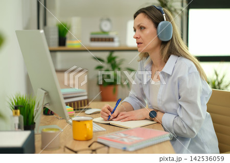 Businesswoman takes notes during online meeting with headphones Businesswoman takes notes during online meeting with headphones 142536059