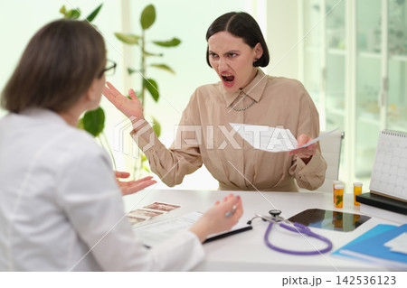 Angry woman arguing with doctor across office desk while holding medical papers in bright clinic interior with stethoscope, test strips, calendar, bottles, and tense consultation mood 142536123