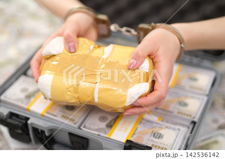 Handcuffed hands holding taped parcel above suitcase filled with bundled cash, suggesting illegal exchange, evidence seizure, contraband transport, fraud investigation, and financial crime concept 142536142