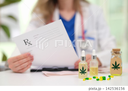 Medical professional holding prescription beside cannabis oil bottles and capsules on desk, suggesting alternative healthcare consultation, pharmacy treatment, wellness guidance, dosage planning Medical professional holding prescription beside cannabis oil bottles and capsules on desk, suggesting alternative healthcare consultation, pharmacy treatment, wellness guidance, dosage planning 142536156