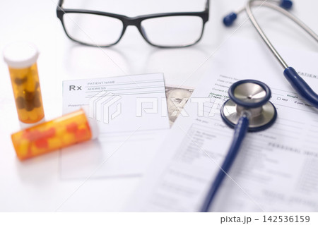 Prescription paperwork with stethoscope, eyeglasses, pill bottles and medical forms on bright white desk in healthcare office, showing medicine management, bribery and insurance administration Prescription paperwork with stethoscope, eyeglasses, pill bottles and medical forms on bright white desk in healthcare office, showing medicine management, bribery and insurance administration 142536159