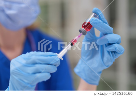 Gloved medical worker drawing red liquid into syringe from small vial during laboratory procedure, close up view with soft focus background and clinical blue uniform Gloved medical worker drawing red liquid into syringe from small vial during laboratory procedure, close up view with soft focus background and clinical blue uniform 142536160