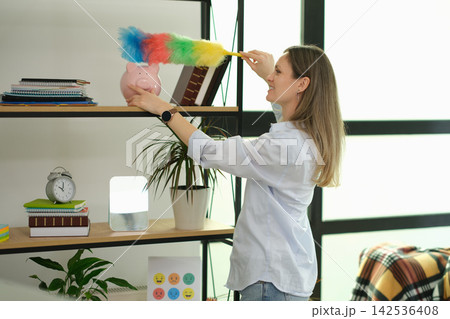 Businesswoman dusts office shelf while holding piggy bank 142536408