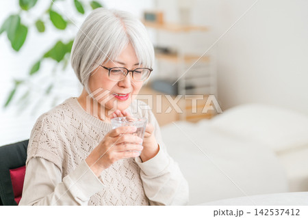 自宅で水を飲むシニア女性。服薬と健康管理をする日常生活 自宅で水を飲むシニア女性。服薬と健康管理をする日常生活 142537412